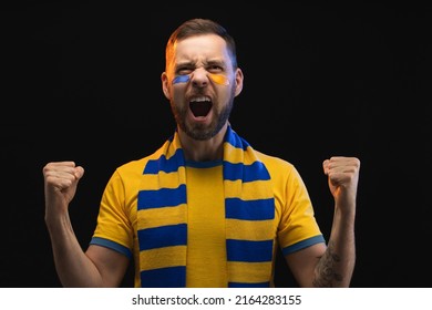 Portrait of euphoric happy soccer supporter man in yellow-blue t-shirt and scarf and painted face, cheering for his favourite team, isolated over black background. - Shutterstock ID 2164283155