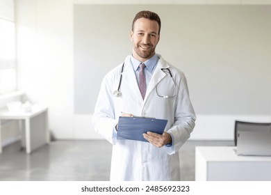 Portrait of European man doctor in work clothes with a stethoscope around his neck, posing with clipboard in hands in clinic, looking and smiling at camera - Powered by Shutterstock