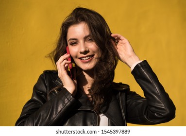 Portrait Of European Young Beautiful Smiling Woman With Dark Straight Hair In Black Leather Jacket Talking To Phone On Yellow Wall Background