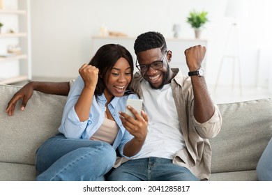 Portrait Of Excited African American Couple Using Smartphone, Young Lady And Guy Making Winner Sign Gesture, Shaking Fists, Sitting On Couch In Living Room, Playing Video Games Or Reading Great News