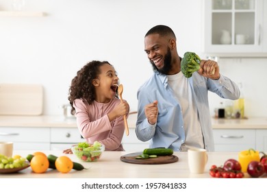 Portrait of excited African American father and his little daughter singing and dancing while cooking together at the kitchen, using spatula and broccoli as microphones, looking at each other - Powered by Shutterstock