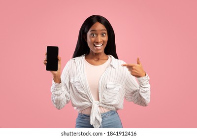 Portrait of excited African American woman pointing at smartphone with empty screen on pink background, mockup for mobile app design. Happy black lady holding cellphone with space for website