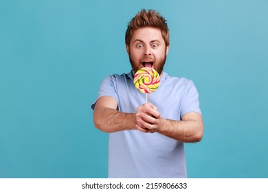 Portrait of excited amazed hungry handsome bearded man holding out sweet sugary confectionery, feels hungry, enjoying tasty lollipop. Indoor studio shot isolated on blue background.