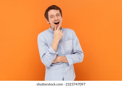 Portrait of excited amazed young adult man standing with hand on chin, having good idea, looking at camera, wearing light blue shirt. Indoor studio shot isolated on orange background.