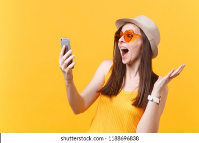 Portrait of excited amazed young woman in straw summer hat, orange glasses keeping mouth wide open, looking surprised, hold mobile phone isolated on yellow background. People sincere emotions concept