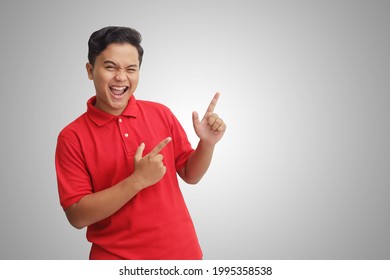 Portrait of excited Asian man in red polo shirt smiling and looking at the camera pointing with two hands and fingers to the side. Isolated image on gray background