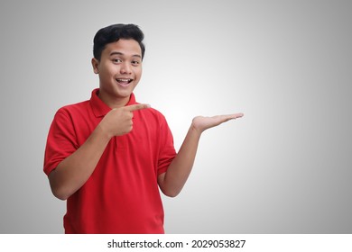 Portrait Of Excited Asian Man In Red Polo Shirt Smiling And Looking At The Camera Showing Something With Hands. Isolated Image On Gray Background