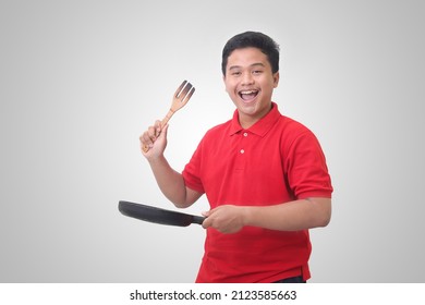 Portrait Of Excited Asian Man In Red Polo Shirt Holding Pan And Spatula, Cooking A Food. Isolated Image On White Background