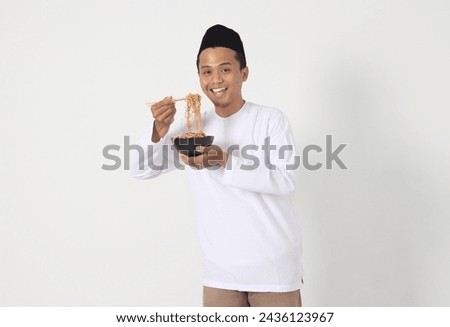 Portrait of excited Asian muslim man eating delicious instant noodles with chopsticks served on plate. Pre dawn meal and break fasting concept. Isolated image on white background