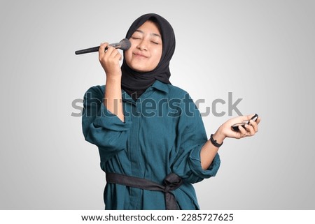 Portrait of excited Asian muslim woman with hijab, applying foundation powder or blush with makeup brush on her cheek. Isolated image on white background