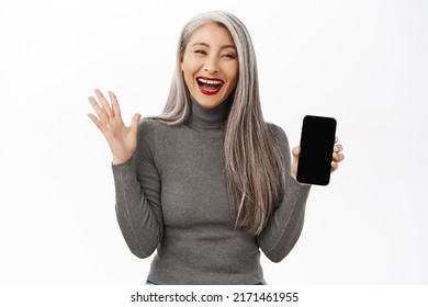 Portrait Of Excited Asian Senior Woman Showing Mobile Phone Screen And Celebrating, Achieve Goal On Smartphone App, Standing Happy Against White Background