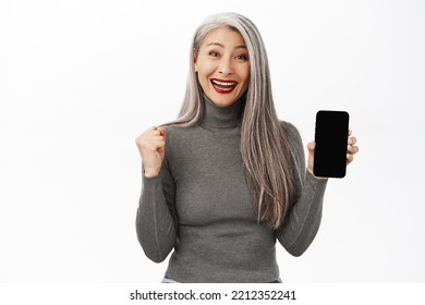 Portrait Of Excited Asian Senior Woman Showing Mobile Phone Screen And Celebrating, Achieve Goal On Smartphone App, Standing Happy Against White Background