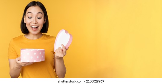 Portrait Of Excited Asian Woman, Open Gift Box With Surprised Happy Face, Standing Over Yellow Background