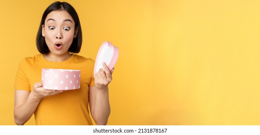 Portrait Of Excited Asian Woman, Open Gift Box With Surprised Happy Face, Standing Over Yellow Background