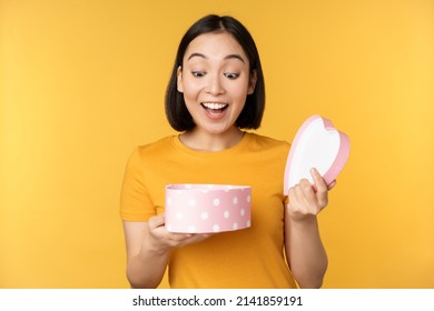 Portrait Of Excited Asian Woman, Open Gift Box With Surprised Happy Face, Standing Over Yellow Background