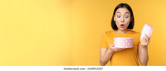 Portrait Of Excited Asian Woman, Open Gift Box With Surprised Happy Face, Standing Over Yellow Background