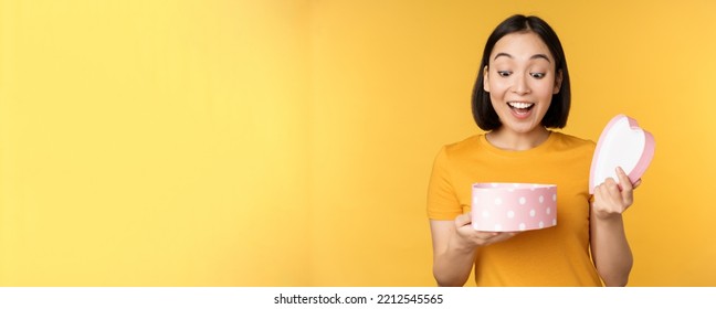 Portrait Of Excited Asian Woman, Open Gift Box With Surprised Happy Face, Standing Over Yellow Background