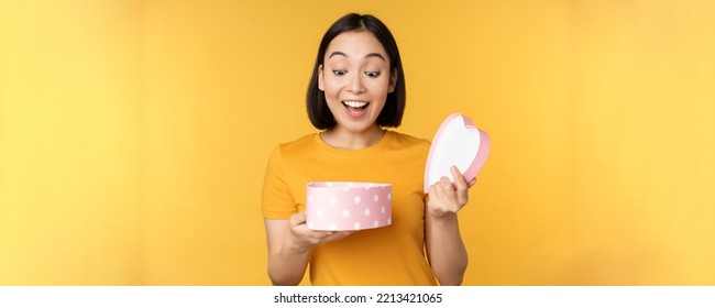 Portrait Of Excited Asian Woman, Open Gift Box With Surprised Happy Face, Standing Over Yellow Background