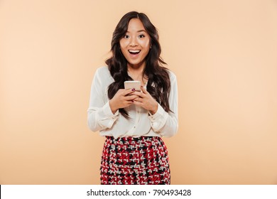 Portrait Of An Excited Asian Woman Standing And Using Mobile Phone Isolated Over Beige Background