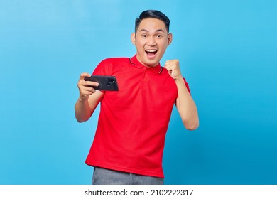 Portrait Of Excited Asian Young Man Playing Game On Mobile Phone And Doing Celebration Gesture For Winning Game Isolated On Blue Background