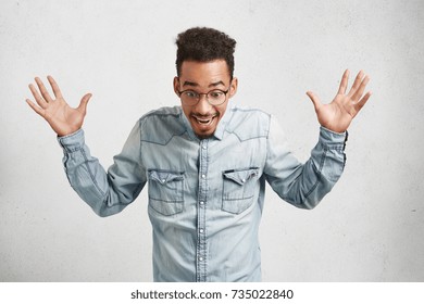 Portrait of excited bearded African American male wears casual denim shirt and round spectacles, gestures actively, looks with great surpisment down, sees unexpected surprise prepared by friends