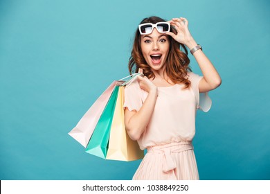 Portrait of an excited beautiful girl wearing dress and sunglasses holding shopping bags isolated over blue background - Shutterstock ID 1038849250