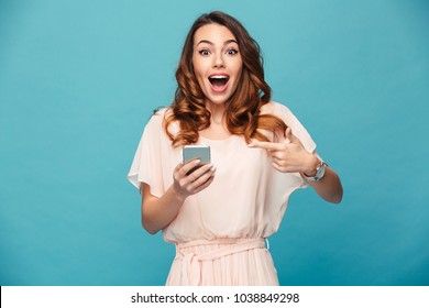 Portrait of an excited beautiful girl wearing dress pointing finger at mobile phone isolated over blue background
