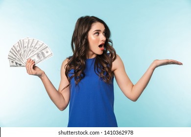 Portrait Of An Excited Beautiful Young Brunette Woman Standing Isolated Over Blue Background, Showing Money Banknotes