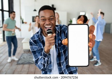 Portrait Of Excited Black Guy Holding Mic And Big Smartphone With White Blank Screen In Hand, Showing Device Close To Camera. Diverse Group Of Friends Singing Using Karaoke App, Free Copy Space