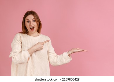 Portrait of excited blond woman presenting copy space for advertisement or promotional text, looking at camera with open mouth, wearing white sweater. Indoor studio shot isolated on pink background.