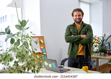 Portrait Of Excited Caucasian Manager Designer Man In Office Posing At Camera, Standing Behind Desk With Laptop, Many Plants In Office Room. European Guy In Casual Wear, Success And Business