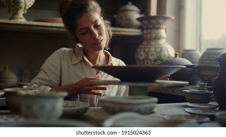 Portrait of excited ceramist examining clay product in workshop. Closeup young woman spinning potters wheel in studio. Smiling female artist enjoying plate in pottery. - Powered by Shutterstock