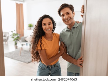 Portrait of excited cheerful couple inviting guest friends to enter their home, happy young guy and lady standing in doorway of contemporary flat, millennial family holding door looking out together - Powered by Shutterstock