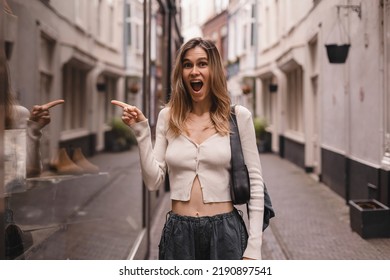 Portrait of excited cheerful female shopaholic standing by showcase and saying wow looking at camera, point her finger on window of shop with shoes and clothes. Discount. Fashion style girl on street.