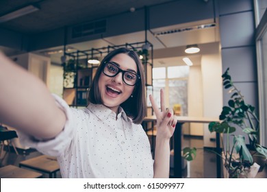 Portrait of excited cheerful smiling young pretty woman in spectacles making selfie photo and showing v-sign with two fingers