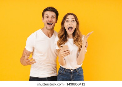 Portrait of excited couple man and woman in basic t-shirts rejoicing while standing together with smartphone isolated over yellow background