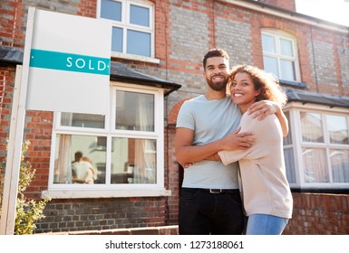 Portrait Of Excited Couple Standing Outside New Home With Sold Sign