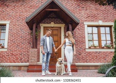 Portrait Of Excited Couple Standing Outside New Home With Dog. Happy Smiling Family Looking At The Labrador In Front Of The House.