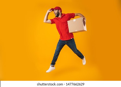 Portrait of excited delivery man in red uniform running and holding parcel box isolated over yellow background
