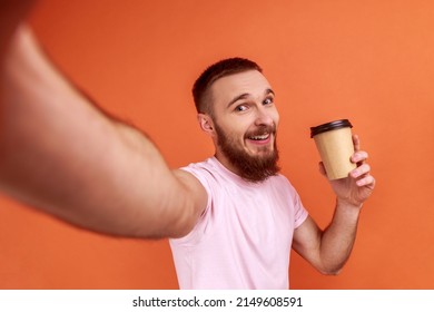 Portrait Of Excited Energetic Bearded Man Taking Selfie With Disposable Cup Of Coffee, POV, Looking At Camera With Optimism, Wearing Pink T-shirt. Indoor Studio Shot Isolated On Orange Background.