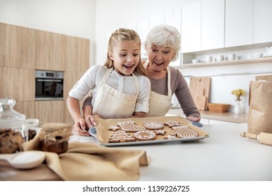 Portrait of excited girl looking at self-made cookies with joy. Her granny is holding a tray and embracing kid with proud. Family is laughing