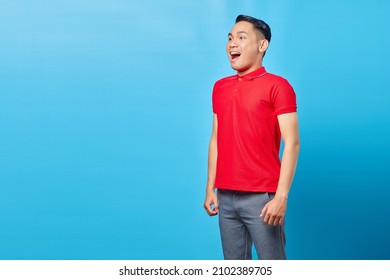 Portrait Of Excited Handsome Asian Young Man In Red Shirt Shouting Sideways With Smiling Facial Expression Isolated On Blue Background