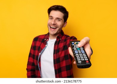 Portrait Of Excited Handsome Guy Watching TV Show Or Film, Holding Remote Control, Switching Channels, Selective Focus On Hand. Young Smiling Man Spending Weekend Free Time, Yellow Studio Background