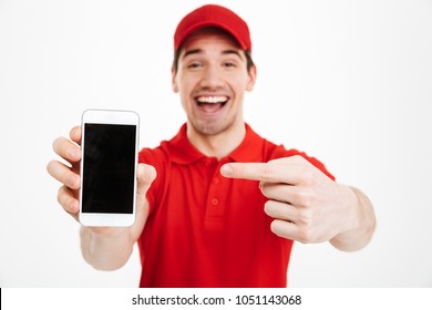 Portrait Of A Excited Happy Young Delivery Man In Red Cap Standing Isolated Over White Background. Looking Camera Showing Display Of Mobile Phone.