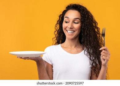 Portrait of excited hungry young lady holding and showing fork and empty plate in hand, funny young woman wants to eat posing isolated on yellow orange studio background, free copy space, mockup place