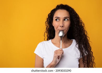 Portrait Of Excited Hungry Young Woman Holding Spoon In Her Mouth Dreaming About Delicious Healthy Food, Choosing Something To Eat Looking Aside At Free Copy Space, Yellow Orange Studio Background