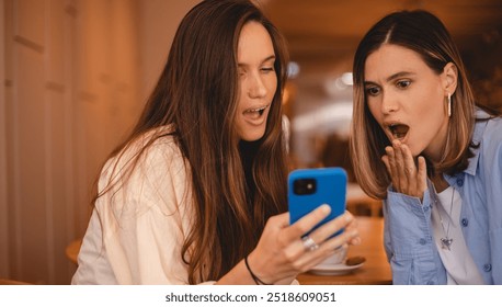 Portrait of excited impressed two  women sisters friends fellows use devices recommend select choose decide discount options, point finger on phone, screaming wow, open mouth, omg, sitting in cafe. - Powered by Shutterstock