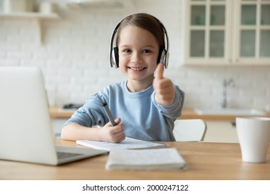 Portrait of excited little 7s Caucasian girl child in headphones show thumb up recommend distant education course. Smiling small kid in earphones give recommendation to online school or webcam class.