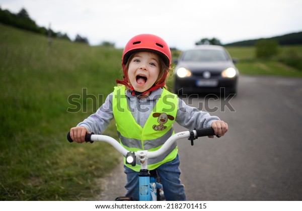 Portrait\
of excited little girl in reflective vest riding bike on road with\
car behind her, road safety education\
concept.