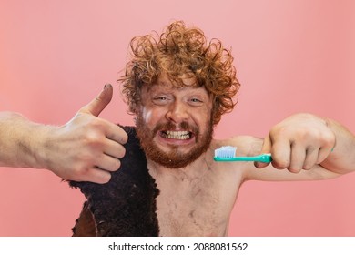 Portrait Of Excited Man In Character Of Neanderthal Brushing Teeth With Toothpaste Isolated Over Pink Background. Like Gesture. Concept Of History, Modern And Ancient Combination, Evolution And Ad